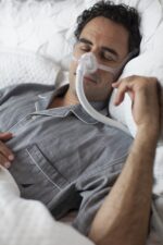 Man sleeping in bed wearing gray pajamas and a CPAP mask for sleep apnea therapy.