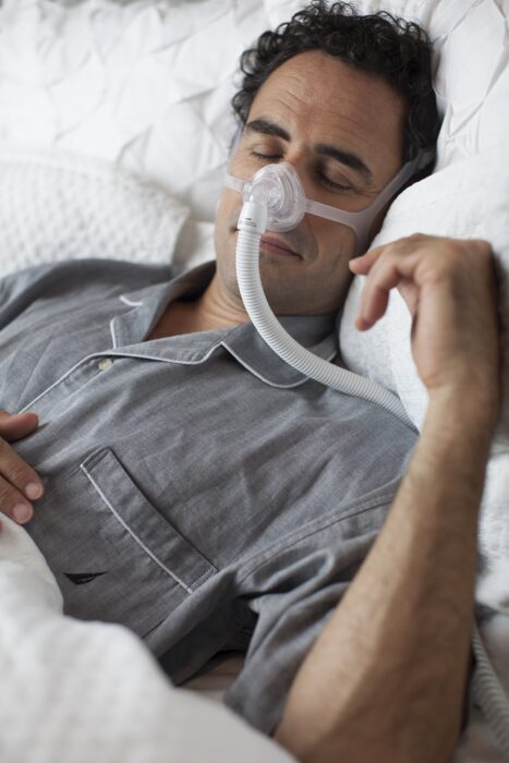 Man sleeping in bed wearing gray pajamas and a CPAP mask for sleep apnea therapy.