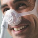 Smiling person wearing a clear nasal CPAP mask with headgear and attached breathing tube.