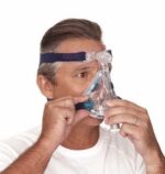 Man wearing and adjusting a clear CPAP face mask with head straps against a white background.