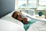 Man sleeping in bed with a CPAP mask and machine, used for sleep apnea therapy, by a window.