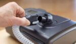 A hand connects a hose to a CPAP machine on a wooden surface.