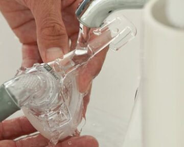 Hands washing a CPAP mask under running water from a faucet near a white sink.