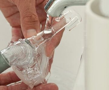 Hands washing a CPAP mask under running water from a faucet near a white sink.