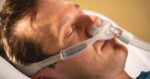 Man lying in bed with eyes closed, wearing a nasal oxygen mask for respiratory support.