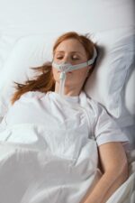 A woman lies in a hospital bed wearing a breathing mask, covered with a white blanket.