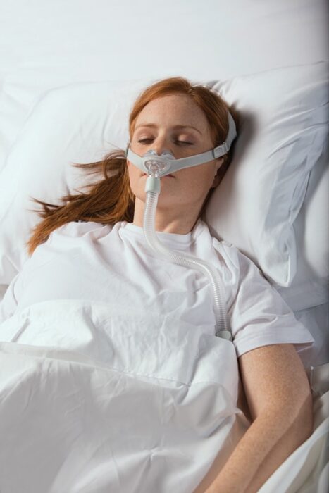 A woman lies in a hospital bed wearing a breathing mask, covered with a white blanket.