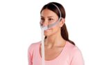 Woman wearing a nasal CPAP mask with headgear and hose, wearing a pink shirt, facing slightly to the side.