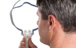 Man holding a nasal CPAP mask near his nose, seen from behind against a white background.