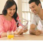 A woman and man smile while examining a CPAP mask at a table with a glass of orange juice.