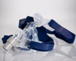 Clear plastic CPAP mask with blue straps placed on a white reflective surface.