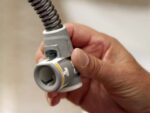 A hand holding a gray plastic connector with a flexible metal hose attached.