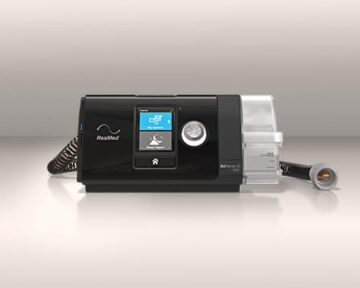 Black ResMed CPAP machine with display screen, hose, and attached clear water chamber on a neutral background.