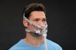 Man wearing a clear CPAP mask with head straps and a tube, against a plain dark background.