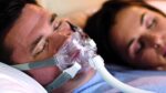 Man sleeping with a CPAP mask beside a woman, both lying in bed.