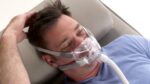 A man lies on a bed wearing a CPAP mask and blue shirt, with one hand touching his head.