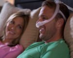 Man wearing a CPAP mask in bed, smiling, with a woman next to him also smiling.