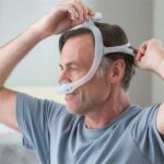 Man putting on a CPAP nasal mask headgear, wearing a light blue shirt, indoors.