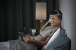 Man in bed wearing a CPAP mask, reading a tablet, with a lamp and CPAP machine on the nightstand.