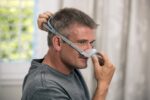 Man putting on a CPAP nasal mask, adjusting the head strap in a bright room.