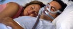 Man sleeping with a CPAP mask next to a woman in bed, both resting peacefully.
