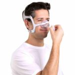 Man wearing a CPAP nasal mask and holding a detachable nasal cushion, isolated on a white background.