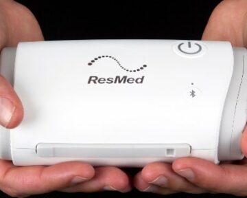Two hands holding a small white ResMed medical device with a power button and Bluetooth symbol.
