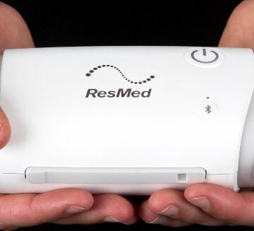 Two hands holding a small white ResMed medical device with a power button and Bluetooth symbol.