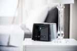 Black CPAP machine and a clear lamp on a white nightstand in a bright bedroom setting.