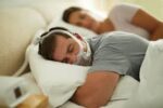 Man sleeping with a CPAP mask on, lying on his side, next to a woman in bed.