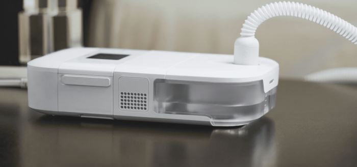 A Phillips Respironics Dreamstation Go Heated Humidifier is sitting on a table next to a bed.