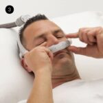 A man is wearing a ResMed AirFit P30i Nasal Pillow CPAP Mask while in bed.