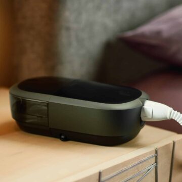 A Respironics Dreamstation 2 Auto CPAP Advanced with Humidifier, Cell Modem & Standard Tubing is sitting on a table next to a bed.