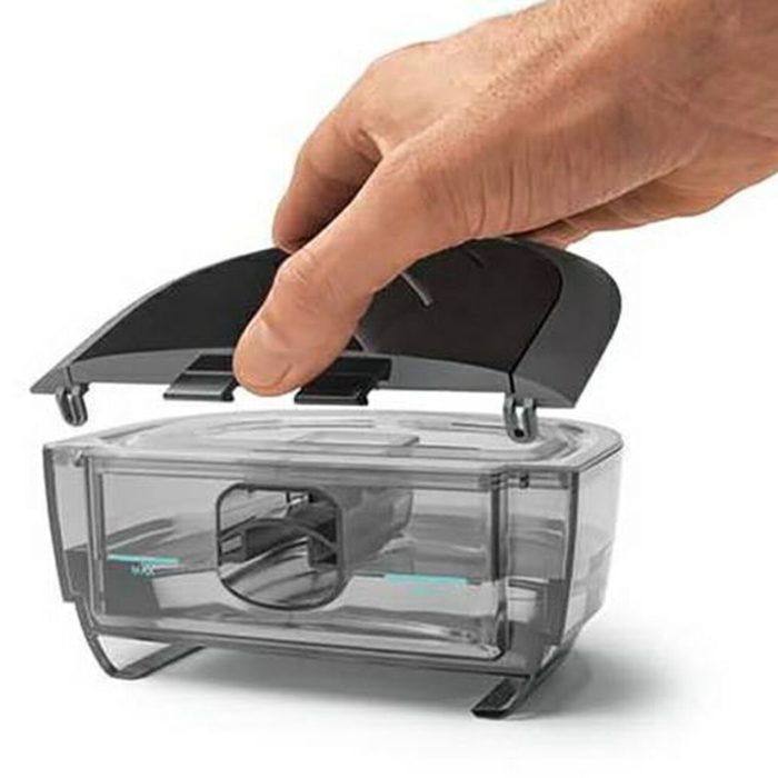 A hand is holding a plastic container with a lid on it, containing the Respironics Dreamstation 2 Humidifier Chamber.