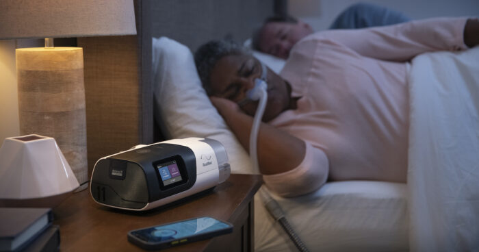 A woman is laying in bed next to a ResMed AirSense 11 Autoset APAP Machine with HumidAir Humidifier and Heated Tubing.