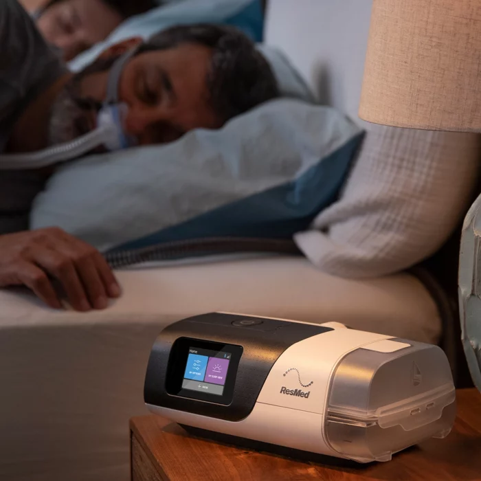 A man is sleeping next to a ResMed AirSense 11 Autoset APAP Machine with HumidAir Humidifier and Heated Tubing.