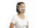 A woman wearing a white t-shirt and a ResMed Airtouch N20 Nasal CPAP mask, smiling slightly, against a white background.