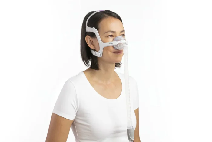 A woman wearing a white t-shirt and a ResMed Airtouch N20 Nasal CPAP mask, smiling slightly, against a white background.