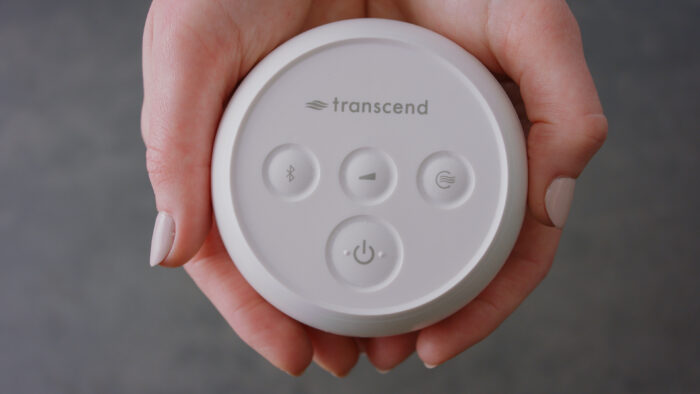Hands holding a white circular device labeled "transcend" with four buttons on its surface, powered by a Transcend PowerAway Micro Battery.