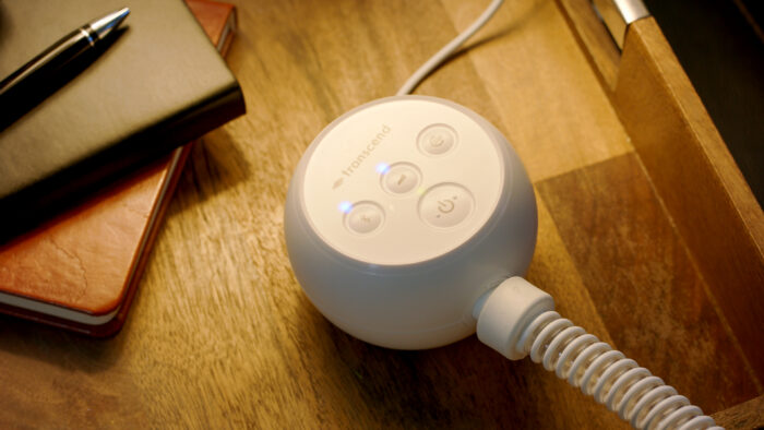 A white Transcend device with control buttons and a coiled cord, powered by a Transcend PowerAway Micro Battery, sits on a wooden surface near a journal and a pen.