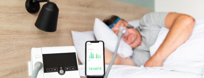 Person with a Lowenstein prismaSMART CPAP/APAP Machine & prismaAQUA Humidifier with 19mm Standard Tubing sleeping in bed, iPhone on bedside table showing sleep data app.