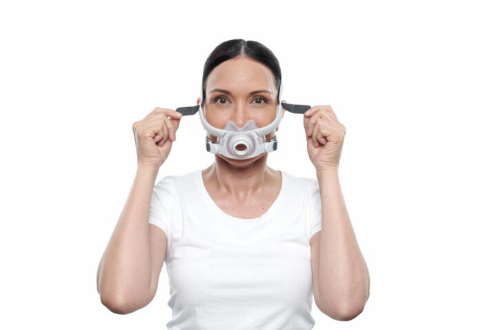 A woman in a white shirt adjusting the straps of her ResMed AirFit F40 Full Face CPAP Mask on her face, isolated on a white background.