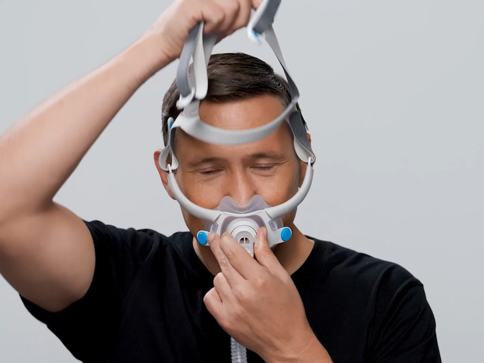 Person wearing a CPAP mask, adjusting the headgear strap over their head against a plain background, creating a sense of routine and comfort at home.