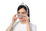 Woman adjusts a ResMed AirTouch N30i Nasal CPAP Mask (Fit Pack) on her face against a white background.