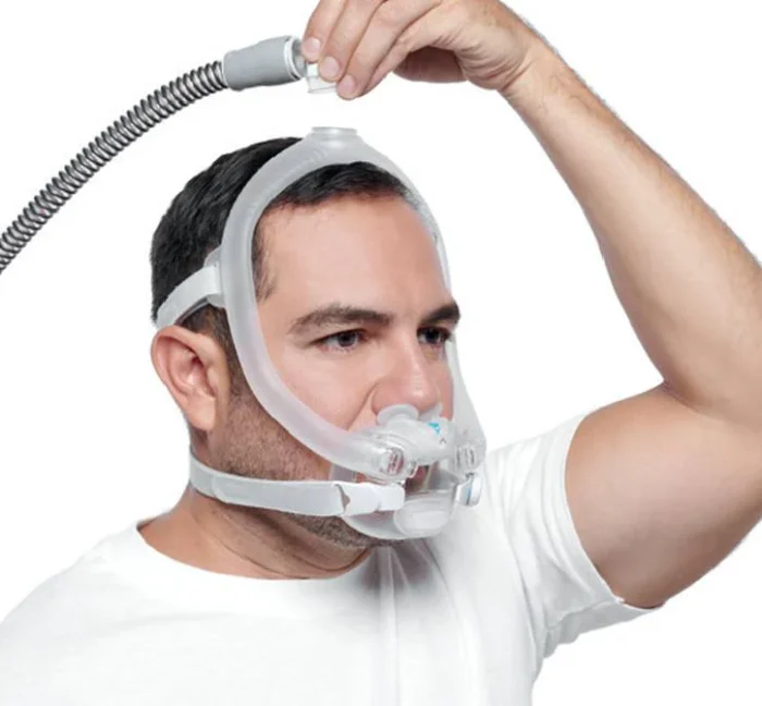 A man connects the hose atop his head while wearing the ResMed AirFit X30i Full Face CPAP Mask, set against a white background.