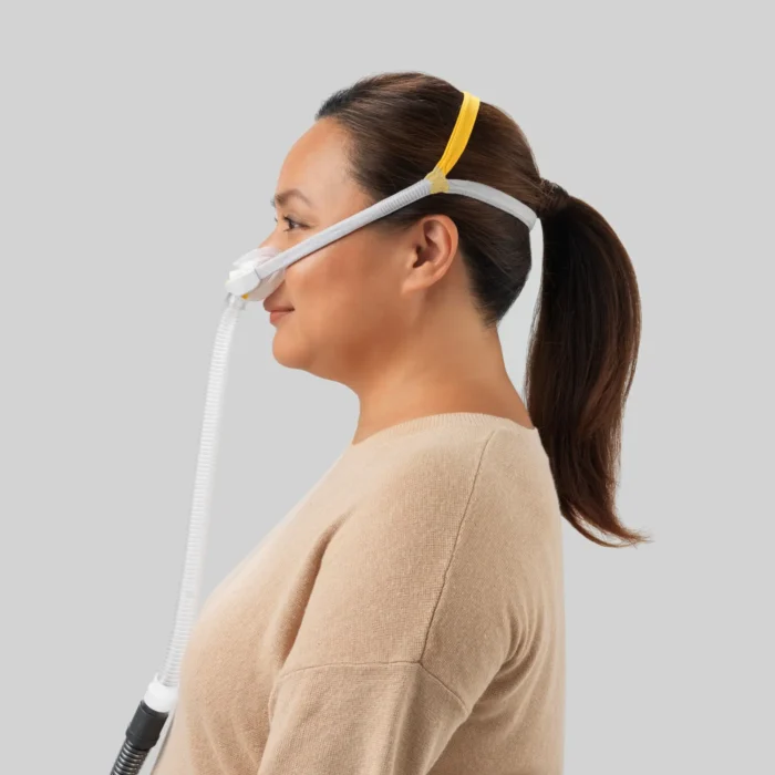 A woman in a beige top wears the Fisher & Paykel Solo Nasal CPAP Mask (Fit Pack) with head straps and hose, shown from the side. The Fit Pack offers personalized comfort for users.