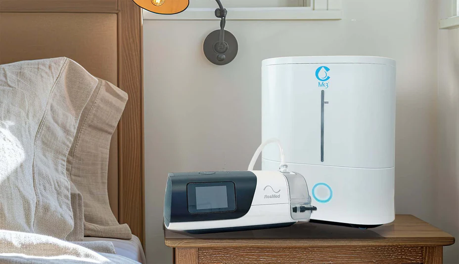 A CPAP machine with a water chamber and an oxygen concentrator sit on a bedside table next to a neatly made bed.