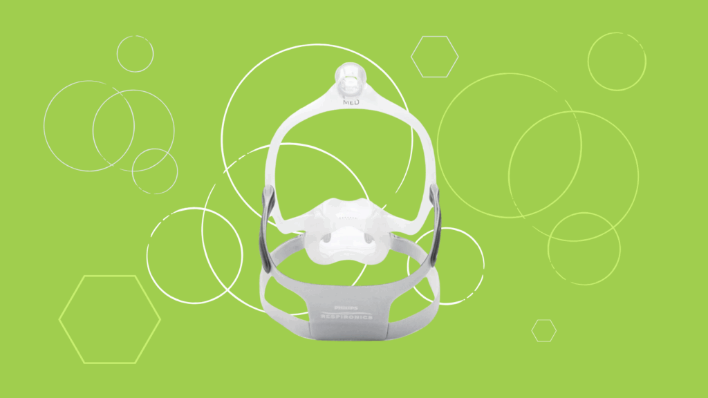 A white Respironics CPAP Mask with gray straps sits on a green background accented by white geometric shapes.